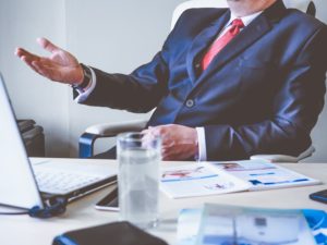 Man in suit sat in meeting room with hand raised from desk